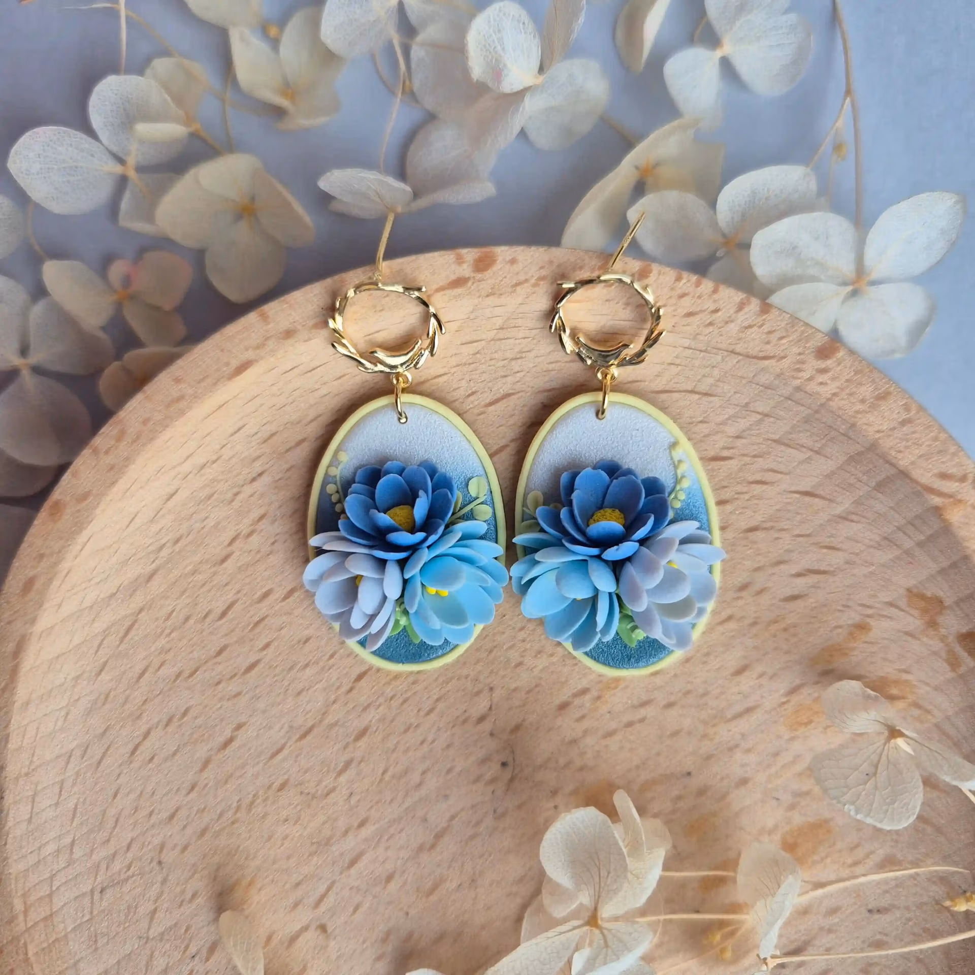 Cercei cu flori 3D modelate din lut polimeric. Florile, in diferite nuante de albastru sunt asezate pe o baza sidefata in degradeu, de la albastru la alb perlat. Cerceii sunt asezati pe un platou de lemn.