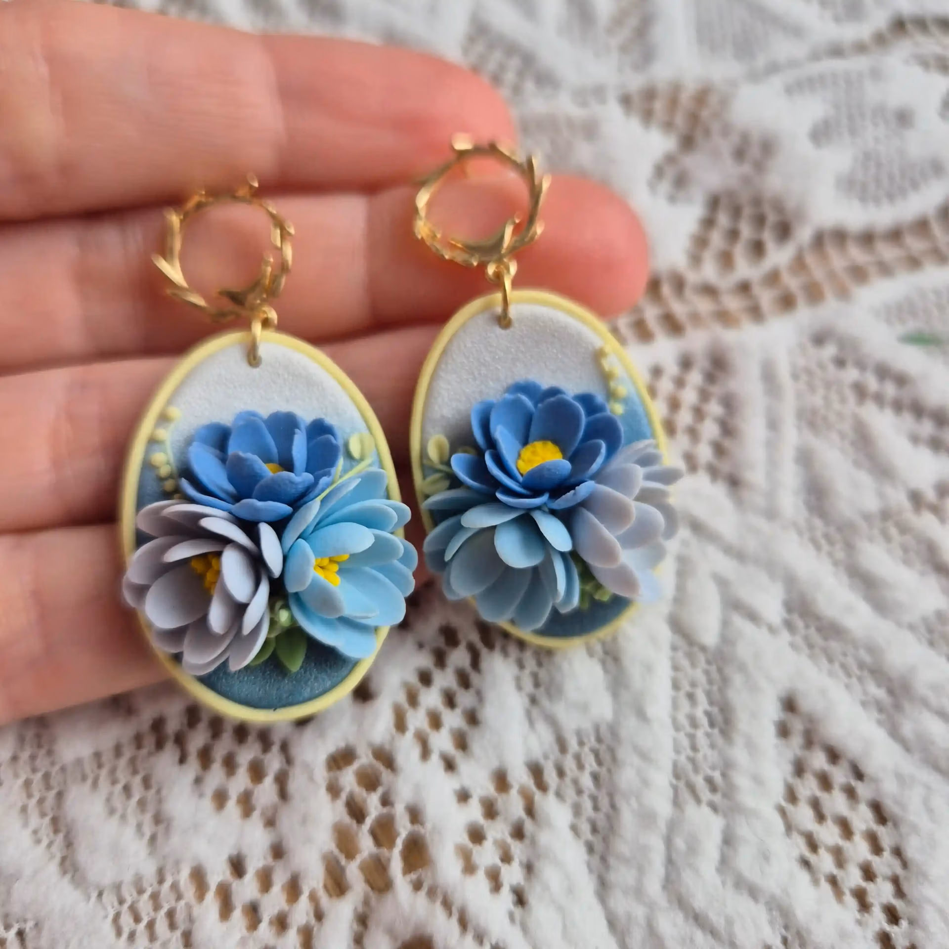 cercei cu flori 3D modelate din lut polimeric. Se poate vedea textura bazei, dar mai ales cat de frumos se vad petalele din lateral. Florile, in diferite nuante de albastru sunt asezate pe o baza sidefata in degradeu, de la albastru la alb perlat. Cerceii sunttinusti in palma, dar decorul general este o bucata de dantela