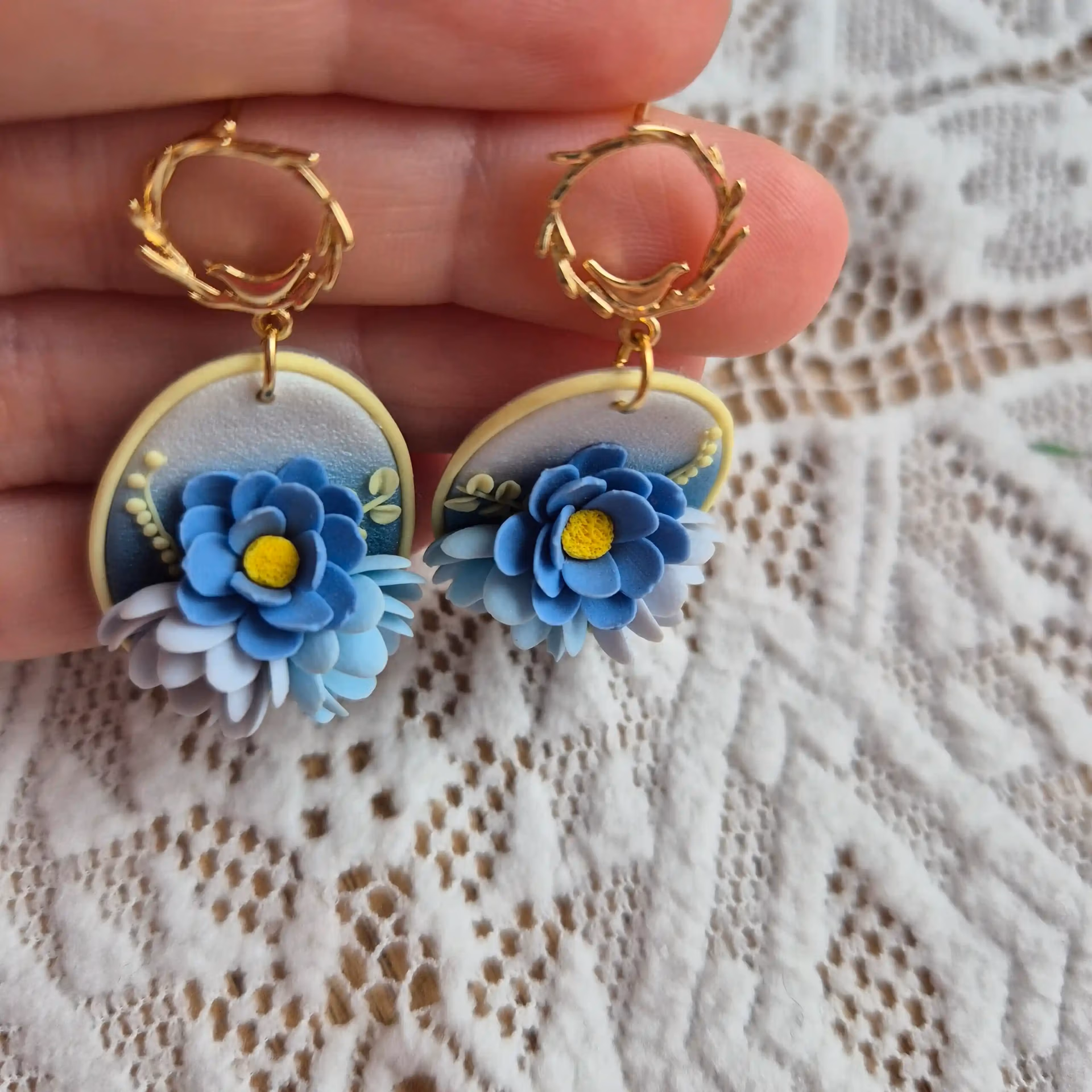 pereche de cercei cu flori 3D modelate din lut polimeric cu accent pus pe una din flori. florile, in diferite nuante de albastru sunt asezate pe o baza sidefata in degradeu, de la albastru la alb perlat. prinderea in ureche este cu o tortita aurie in care se poate vedea o pasare stand pe ramurica