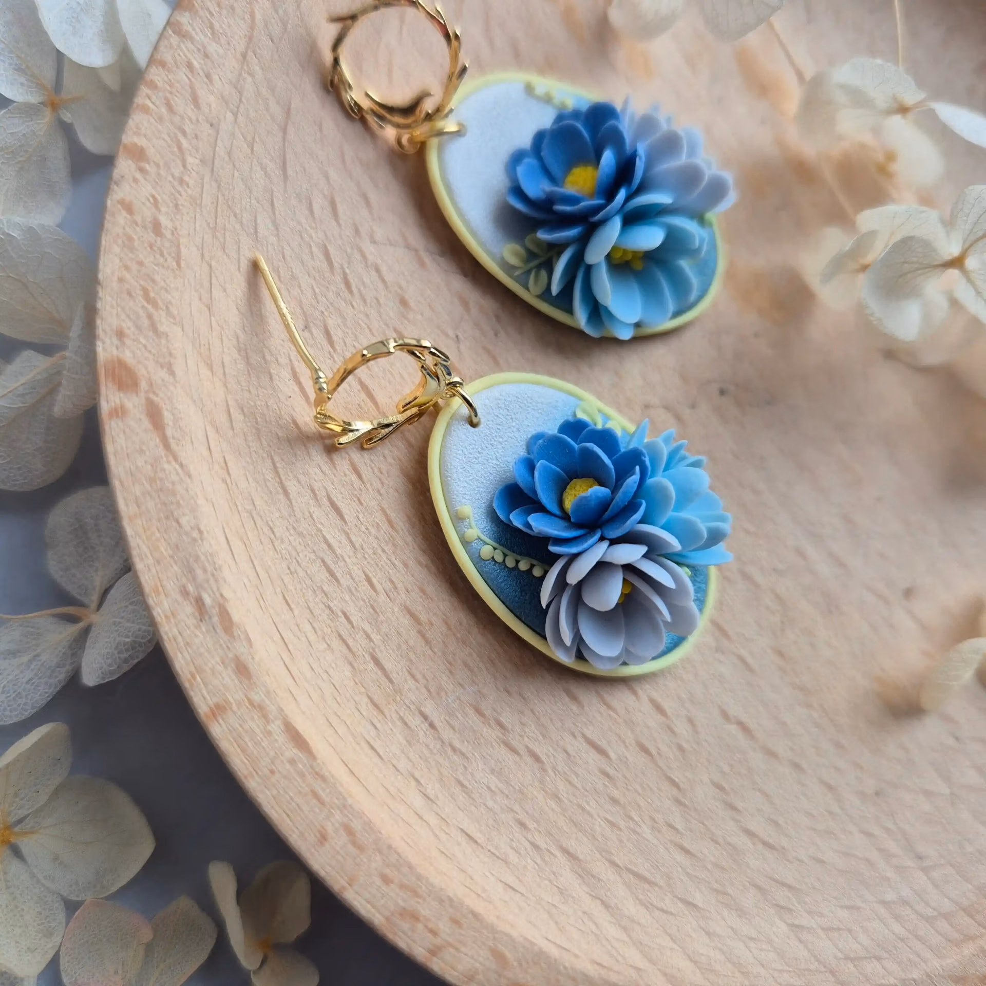 focus pe una din perechile de cercei cu flori 3D modelate din lut polimeric. Se poate vedea textura bazei, dar mai ales cat de frumos se vad petalele din lateral. Florile, in diferite nuante de albastru sunt asezate pe o baza sidefata in degradeu, de la albastru la alb perlat. Cerceii sunt asezati pe un platou de lemn.