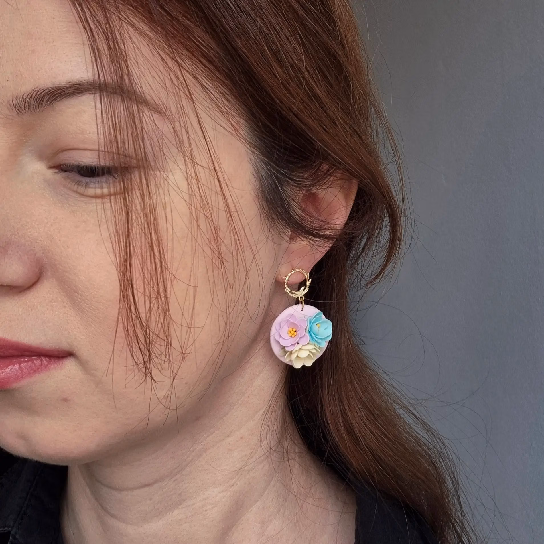 femeie purtand cercei din lut polimeric cu flori modelate 3 D in culori pastelate roz, turcoaz si crem. La una din flori se pot observa staminele galbene. Prinderea pe ureche este in forma de crenguta cu frunze si o pasare