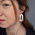 femeie purtand cercei din lut polimeric in forma ovala cu flori dispunse la baza formei. florile, modelate 3 D au culori lavanda, visiniu si galben pai. Prinderea pe ureche este in forma de fundita aurie.