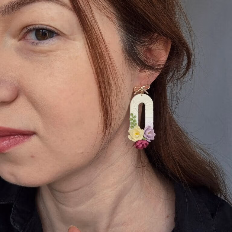 femeie purtand cercei din lut polimeric in forma ovala cu flori dispunse la baza formei. florile, modelate 3 D au culori lavanda, visiniu si galben pai. Prinderea pe ureche este in forma de fundita aurie.
