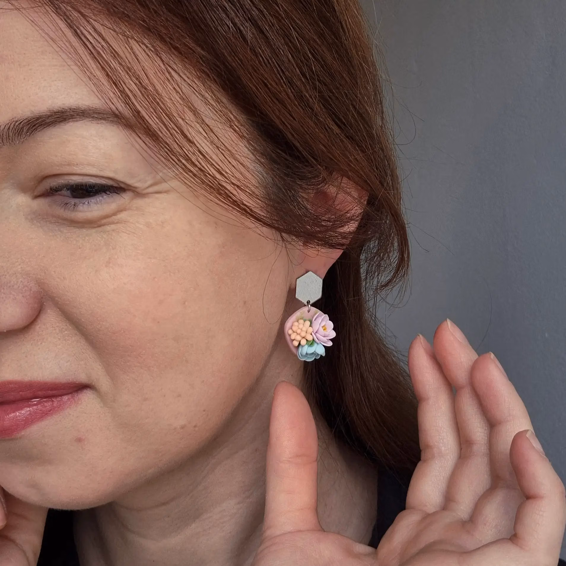 femeie purtand cercei din lut polimeric cu flori modelate 3 D in culori pastelate roz, turcoaz si crem-portocaliu. Prinderea pe ureche este in forma de hexagon argintiu.