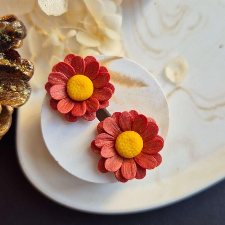 Cercei din lut polimeric reprezentand margarete in culoare rosu-caramiziu. cerceii sunt asezati pe un suport ceramic pentru a se putea observa detaliile