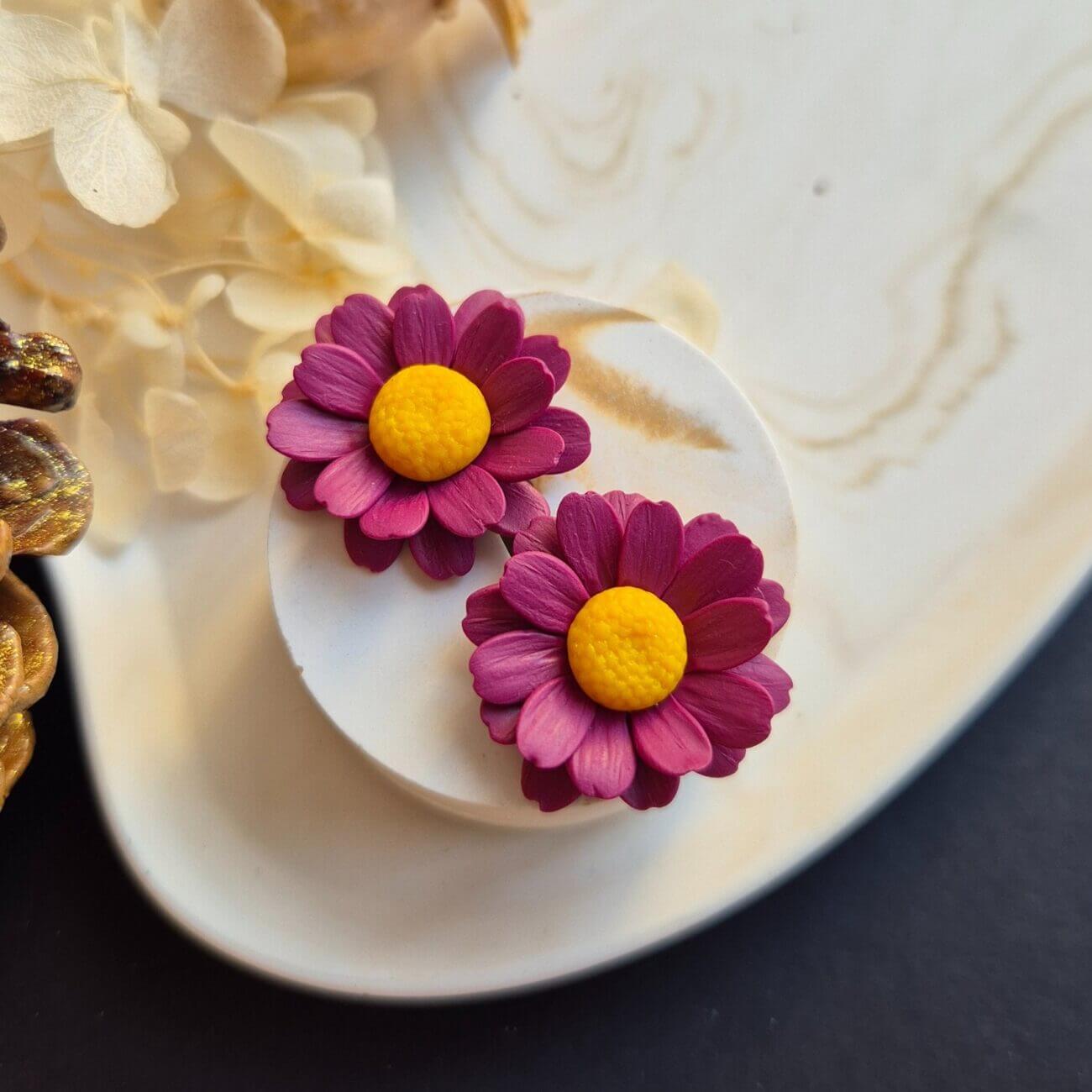 Cercei din lut polimeric reprezentand margarete in culoare burgundy. cerceii sunt asezati pe un suport ceramic deschis la culoare pentru a se vedea usor detaliile si culoarea