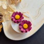 Cercei din lut polimeric reprezentand margarete in culoare burgundy. cerceii sunt asezati pe un suport ceramic deschis la culoare pentru a se vedea usor detaliile si culoarea