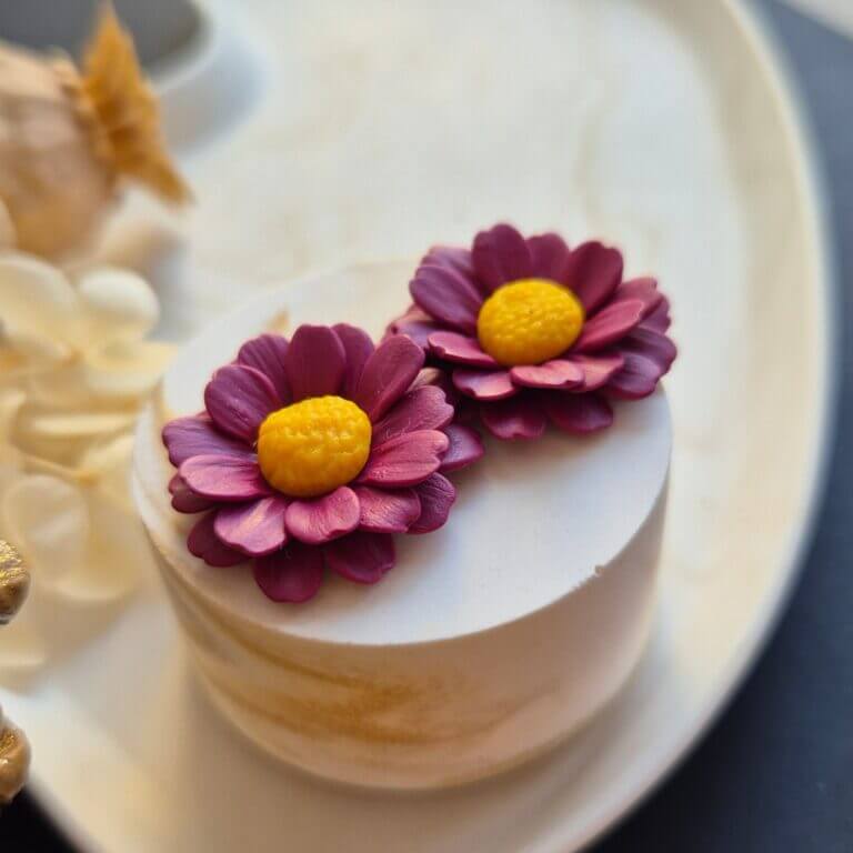 Cercei din lut polimeric reprezentand margarete in culoare burgundy. cerceii sunt asezati pe un suport ceramic pentru a se putea observa detaliile din perspeciva laterala