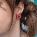 femeie purtand cercei cu fundite create manual din lut polimeric, in culoare rosu caramiziu, sidefat si cu sclipici aplicat local. cerceii sunt purtati pe lobul urechii, tip studs