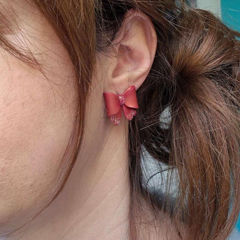 femeie purtand cercei cu fundite create manual din lut polimeric, in culoare rosu caramiziu, sidefat si cu sclipici aplicat local. cerceii sunt purtati pe lobul urechii, tip studs