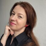 femeie purtand o pereche de cercei in forma de inimioara rosie pe lobul urechii. femeia este zambitoare si pare ca se simte bine purtand acest model de cercei