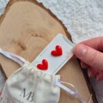 cercei in forma de inimioare rosii din lut polimeric peste care s-a aplicat un strat de rasina pentru extra luciu. Cerceii sunt pentru purtat pe lobul urechii si sunt asezati pe un cartonas personalizat care este introdus partial intr-un saculet textil personalizat