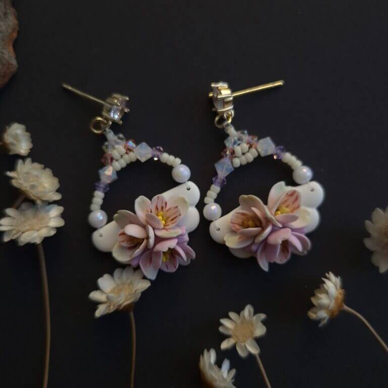 cercei din lut polimeric si cristale, in nuante pale de roz si mov fotografiate pe un fundal negru cu accente de flori uscate
