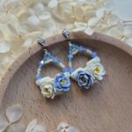 cercei din lut polimeric si cristale, in nuante de albastru si crem-vanilie fotografiate pe un platou de lemn, inconjurat de flori