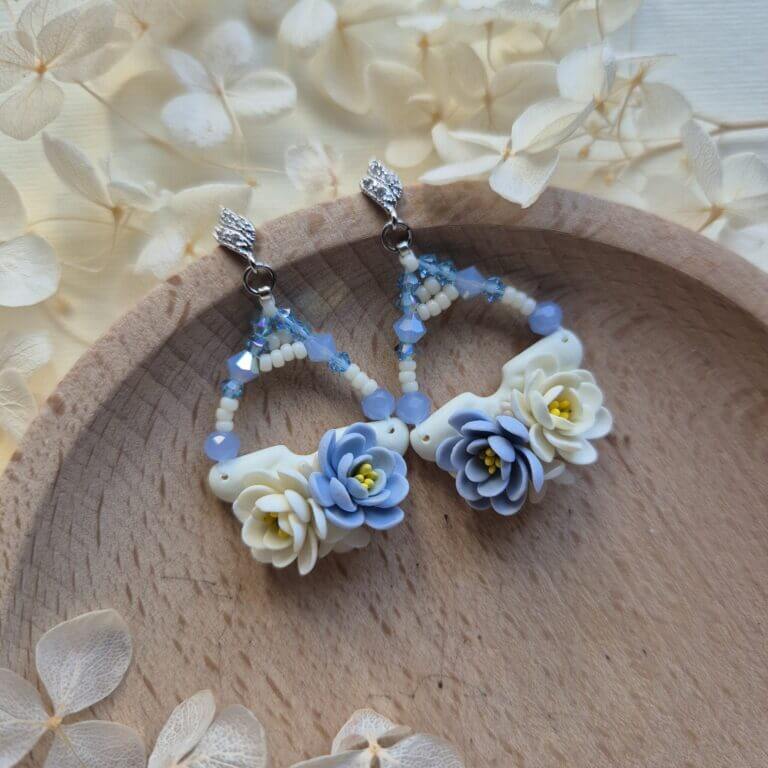 cercei din lut polimeric si cristale, in nuante de albastru si crem-vanilie fotografiate pe un platou de lemn, inconjurat de flori