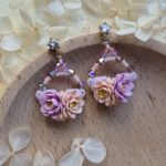 cercei din lut polimeric si cristale, in nuante pale de mov si portocaliu fotografiate pe un platou de lemn, inconjurat de flori