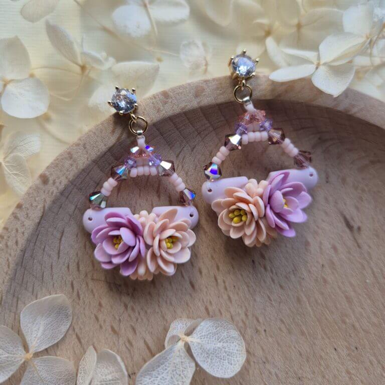 cercei din lut polimeric si cristale, in nuante pale de mov si portocaliu fotografiate pe un platou de lemn, inconjurat de flori