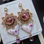 Cercei cu cristale svarovski roz, rivoli in jurul carora sunt cusute margelute de sticla aurii miyuki, partea cu cristale are forma de cerc cu petale zimtate. de aceasta piese este prinsa prin zale aurii o piesa din lut polimeric imprimata cu flori in culori asortate. piesa din lut polimeric este lucioasa pentru ca este lacuita cu rasina UV si are o forma asemanatoare cu un triunghi. in partea de jos sunt prinse cate un cristal roz de sticla in forma de lacrima. Cerceii au tortite cu cristal zirconia si se prin in ureche cu surub si dop de silicon. Cerceii sunt asezati pe un carton texturat personalizat.