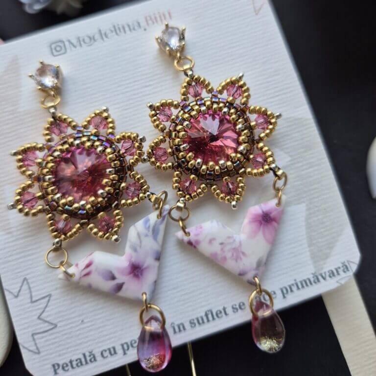 Cercei cu cristale svarovski roz, rivoli in jurul carora sunt cusute margelute de sticla aurii miyuki, partea cu cristale are forma de cerc cu petale zimtate. de aceasta piese este prinsa prin zale aurii o piesa din lut polimeric imprimata cu flori in culori asortate. piesa din lut polimeric este lucioasa pentru ca este lacuita cu rasina UV si are o forma asemanatoare cu un triunghi. in partea de jos sunt prinse cate un cristal roz de sticla in forma de lacrima. Cerceii au tortite cu cristal zirconia si se prin in ureche cu surub si dop de silicon. Cerceii sunt asezati pe un carton texturat personalizat.