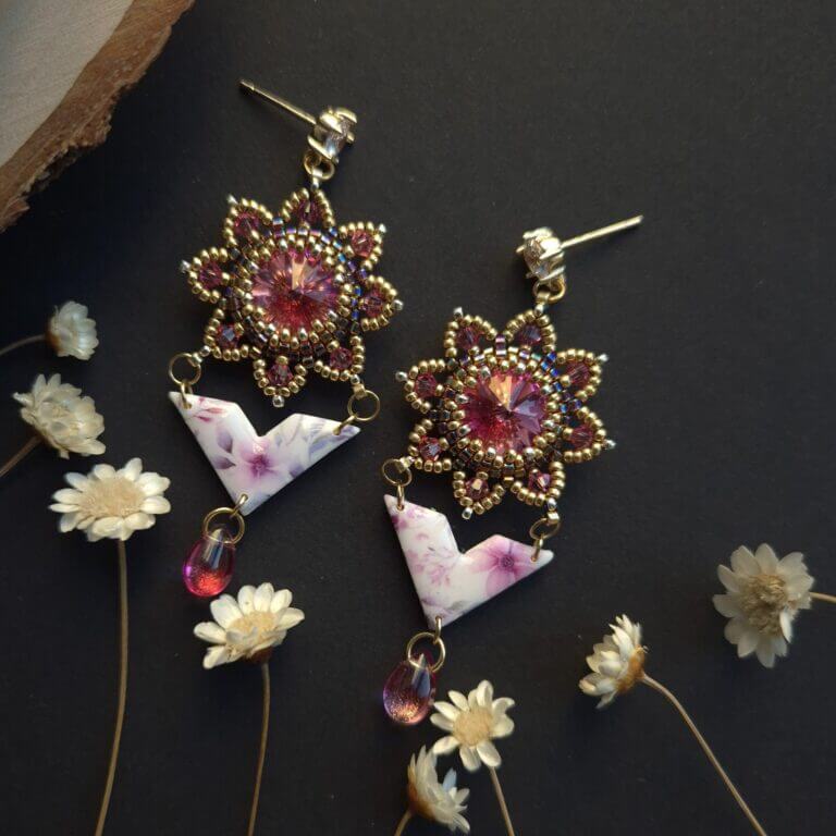 Cercei cu cristale svarovski roz, rivoli in jurul carora sunt cusute margelute de sticla aurii miyuki, partea cu cristale are forma de cerc cu petale zimtate. de aceasta piese este prinsa prin zale aurii o piesa din lut polimeric imprimata cu flori in culori asortate. piesa din lut polimeric este lucioasa pentru ca este lacuita cu rasina UV si are o forma asemanatoare cu un triunghi. in partea de jos sunt prinse cate un cristal roz de sticla in forma de lacrima. Cerceii au tortite cu cristal zirconia si se prin in ureche cu surub si dop de silicon. Cerceii sunt fotografiati pe un fundal negru si se vad in partea inferioara cateva flori uscate de paie