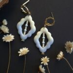 Cercei din lut polimeric cu forma organica in nunate degradee de la albastru la crem. In mijlocul piesei sunt insirate margele din sticla pe un fir metalic. in imagine se vede atentia cu care sunt finisati