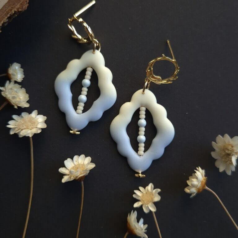 Cercei din lut polimeric cu forma organica in nunate degradee de la albastru la crem. In mijlocul piesei sunt insirate margele din sticla pe un fir metalic. in imagine se vede atentia cu care sunt finisati