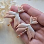 cercei din lut polimeric, catifelati si usori, portocaliu deschis in degrade, avand pe mijloc o piesa mobila cu margele din sticla insirate. cerceii sunt formati din 3 piese legate intre ele cu zale aurii. asezarea in ureche se face cu tortite platou suflate cu aur si sigilate cu un strat de rasina biocompatibila. Partea vizibila a tortitelor are forma de petale. Cerceii sunt tinuti in palma