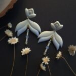 cercei din lut polimeric, catifelati si usori, albastru degrade, avand pe mijloc o piesa mobila cu margele din sticla insirate. cerceii sunt formati din 3 piese legate intre ele cu zale aurii. asezarea in ureche se face cu tortite platou suflate cu aur si sigilate cu un strat de rasina biocompatibila. Partea vizibila a tortitelor are forma de petale. Cerceii sunt fotografiati pe un fundal negru cu decor de flori uscate crem