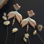 cercei din lut polimeric, catifelati si usori, portocaliu degrade, avand pe mijloc o piesa mobila cu margele din sticla insirate. cerceii sunt formati din 3 piese legate intre ele cu zale aurii. Asezarea in ureche se face cu tortite platou suflate cu aur si sigilate cu un strat de rasina biocompatibila. Partea vizibila a tortitelor are forma de petale. Cerceii sunt fotografiati pe un fundal negru cu decor de flori uscate crem