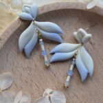 cercei din lut polimeric, catifelati si usori, albastru deschis in degrade, avand pe mijloc o piesa mobila cu margele din sticla insirate. cerceii sunt formati din 3 piese legate intre ele cu zale aurii. asezarea in ureche se face cu tortite platou suflate cu aur si sigilate cu un strat de rasina biocompatibila. Partea vizibila a tortitelor are forma de petale. Cerceii sunt asezati pe un platou de lemn