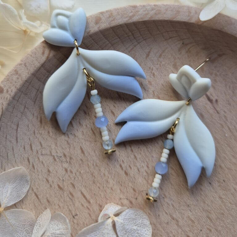 cercei din lut polimeric, catifelati si usori, albastru deschis in degrade, avand pe mijloc o piesa mobila cu margele din sticla insirate. cerceii sunt formati din 3 piese legate intre ele cu zale aurii. asezarea in ureche se face cu tortite platou suflate cu aur si sigilate cu un strat de rasina biocompatibila. Partea vizibila a tortitelor are forma de petale. Cerceii sunt asezati pe un platou de lemn