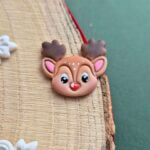 Brosa din lut polimeric reprezentand un ren dragut, cu ochi si nas luciosi, lacuiti cu rasina. detaliile sunt pictate manual. Brosa este asezata pe o felie de lemn