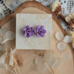 Cercei handmade în formă de floare suculentă mov violet, vedere de sus pe un suport de lemn cu flori uscate albe, design botanic elegant.