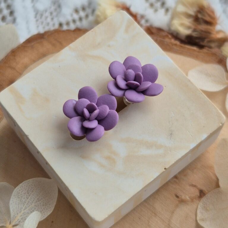 Cercei handmade în formă de floare suculentă mov violet, vedere de sus pe un suport de lemn cu flori uscate albe, design botanic elegant.