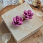 Cercei handmade în formă de floare suculentă mov violet, vedere de sus pe un suport de lemn cu flori uscate albe, design botanic elegant.