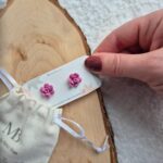 Cercei handmade în formă de floare suculentă mov lavanda, vedere de sus cu o mana tinand un cartonas personalizat pe care sunt asezati cerceii.