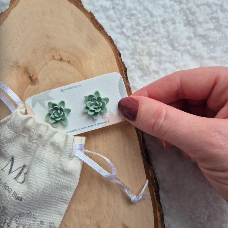 Pereche de cercei tip stud modelați manual în formă de suculentă verde mentă, cu detalii galbene pe vârfurile petalelor, așezați pe un cartonas care are coltul introdus intr-un saculet textil cu care ajunge la tine