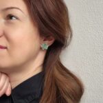 Femeie purtand o pereche de cercei lucrați manual în formă de plante suculente Echeveria, de culoare verde albastru cu detalii galbene pe marginea petalelor.