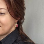 Cercei cu flori magenta,frunze si puncte verzi asezate pe o baza de culoarea piersicii, intr-o forma organica. Cerceii au tortite tip half hoop argintii si sunt asezati purtati de o femeie imbracata in camasa neagra