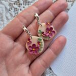 Cercei cu flori magenta,frunze si puncte verzi asezate pe o baza de culoarea piersicii, intr-o forma organica. Cerceii au tortite tip half hoop argintii si sunt tinuti in palma pentru a se putea observa dimeniunile