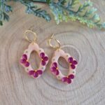 o pereche de cercei din lut polimeric cu margini ondulate, impacabil finisati, avand flori magenta si puncte verzi dispuse pe baza in culoare piersica. piesa cu care se aseaza in ureche este in forma de cerc placat cu aur. Cerceii sunt asezati pe o baza de lemn.