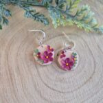 Cercei cu flori magenta,frunze si puncte verzi asezate pe o baza de culoarea piersicii, intr-o forma organica. Cerceii au tortite tip half hoop argintii si sunt asezati pe un platou de lemn si in partea supersioara se observa o frunza