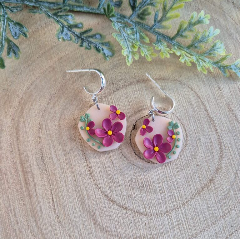 Cercei cu flori magenta,frunze si puncte verzi asezate pe o baza de culoarea piersicii, intr-o forma organica. Cerceii au tortite tip half hoop argintii si sunt asezati pe un platou de lemn si in partea supersioara se observa o frunza